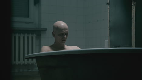 pale bald skinny man takes a bath in the creepy old bathroom at night