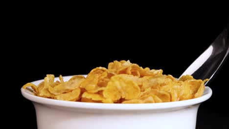 a spoon scoops cornflakes from a white bowl against a black background, highlighting the cereal's texture and color