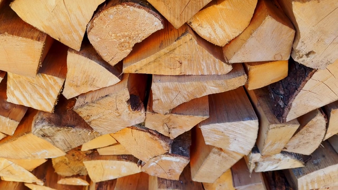Tightly packed split firewood stacked in bright daylight, showing pale cut faces, bark strips, dry log shapes, and rough natural grain texture.