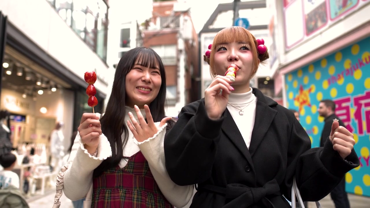 賑やかな通りで、おしゃれな若い女性二人がイチゴとブドウのタンフールー串を楽しむ。
