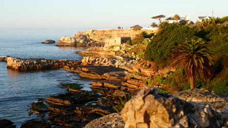 Orange-glow-of-sunrise-on-rugged-coastal-rocks-of-Hermanus-sea-cliffs,-slider