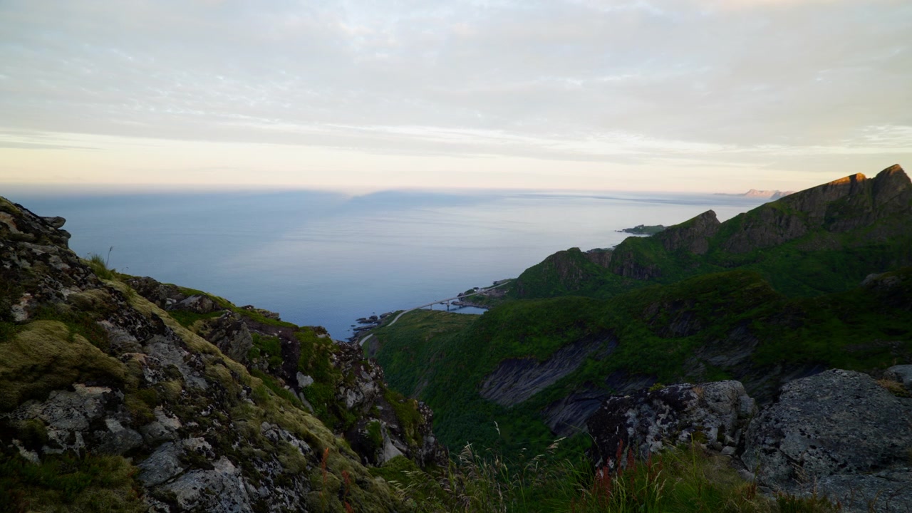 Cahaya lembut pagi hari menyinari tebing pantai berbatu dan laut lepas yang tenang dari lereng gunung yang tinggi di Lofoten, Norwegia.
