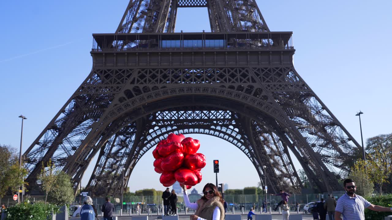 A joyful tourist poses with red heart-shaped balloons by the Eiffel Tower on a sunny day, capturing Paris's iconic romance in slow motion