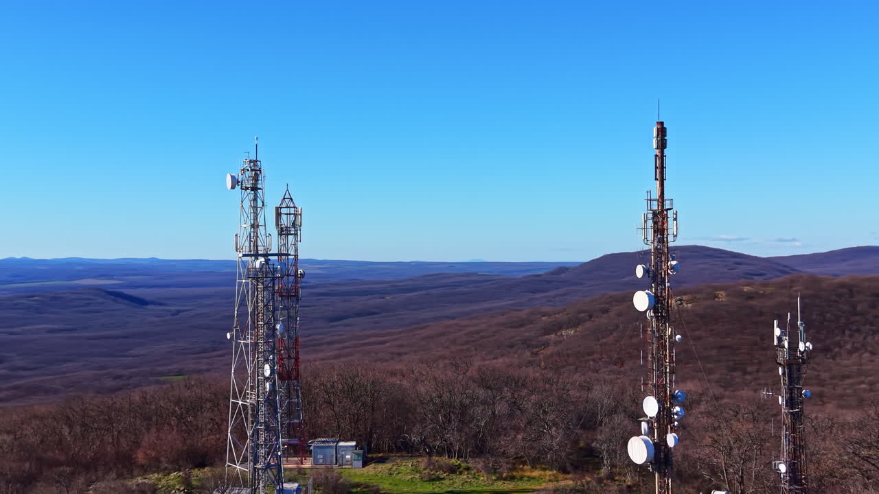 Communication towers stand tall on a mountain overlooking a valley under a clear blue sky in the daytime with no clouds