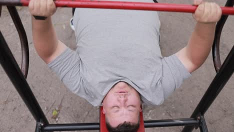 A Fat Man in a Gray Tshirt Lifts Weights while Lying on an Outdoor Exercise Machine a Fat Man in a Gray Tshirt Lifts Weights while Lying on an Outdoor Exercise Machine Struggle with Excess Weight Lifting the Bar