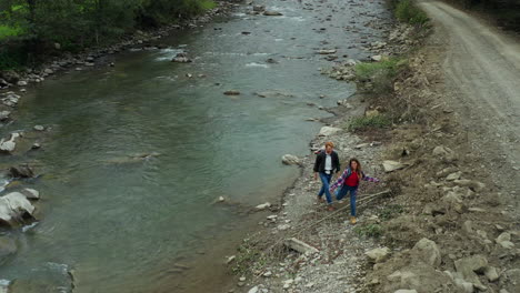 young couple walking through the river shore close to mountain road among green forest trees