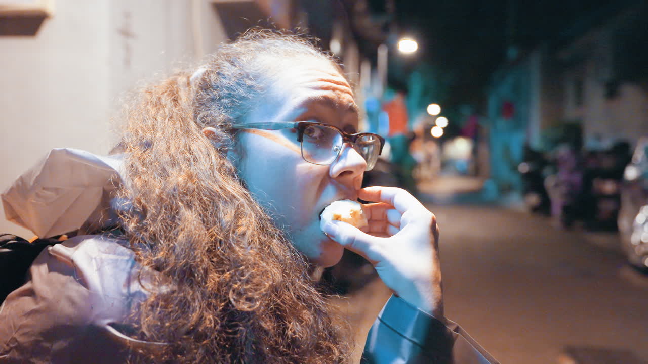Neon alley portrait woman snacking while watching approaching motorcycle, candid profile with glasses, curly hair, warm lamp glow, reflective pavement, quiet tension, urban exploration at night