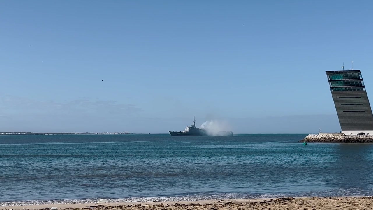 Nave militare che emette fumo vicino alla costa portoghese in una giornata limpida. Una scena marittima suggestiva che mostra attività navale, acque oceaniche calme e una moderna struttura costiera