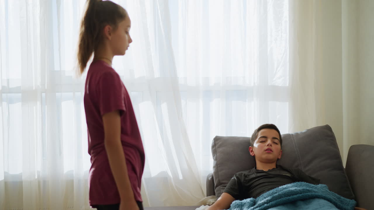 Young boy peacefully sleeping on couch with blue blanket, his sister gently comes to adjust the blanket, showing care and love in a domestic, comforting scene