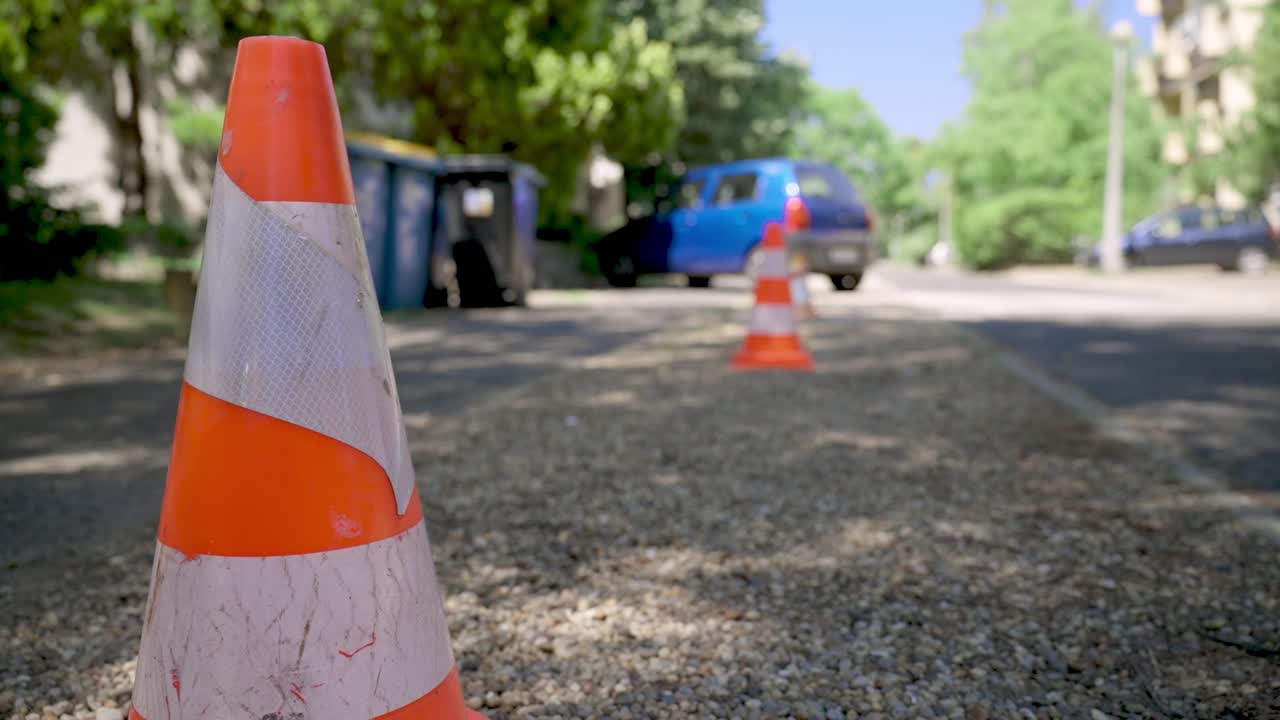 Traffic cones placed on a gravel road near a blue car and garbage bins