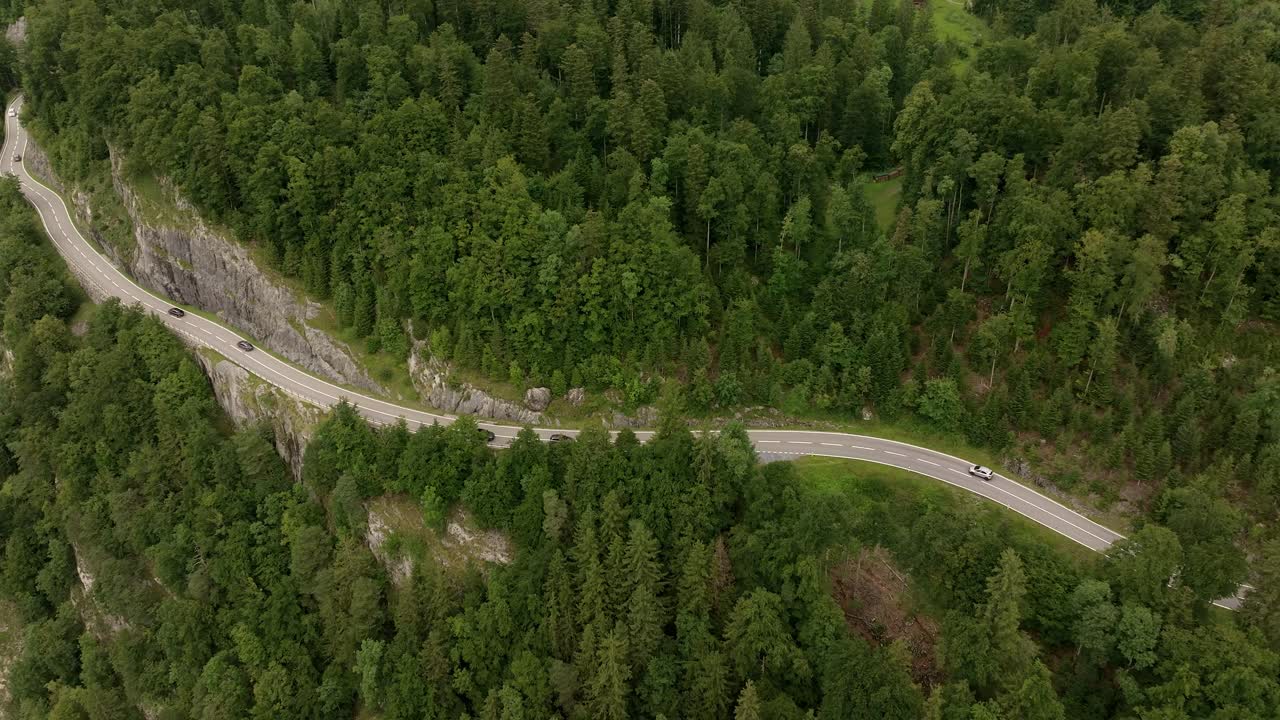Aerial view of a winding mountain road cutting through dense green forest and rocky cliffs. Several cars travel along the scenic route