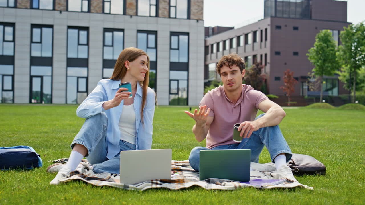 Et par freelancere som sitter i gresset i en bypark og nyter kaffe. Smilende mann og kvinne