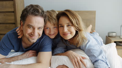 close-up view of happy father and mother with his cute little son hugging while they lying on the bed and looking at camera in the morning