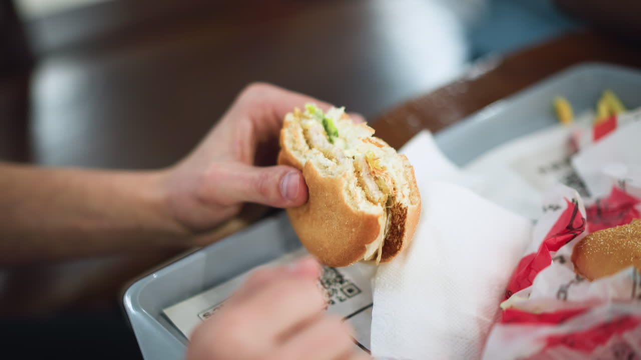 Half eaten burger held in hand closeup showing bite missing with sauce, crumbs on fingers, blurred background suggesting casual dining moment, grease shine and texture conveying hunger and satisfaction