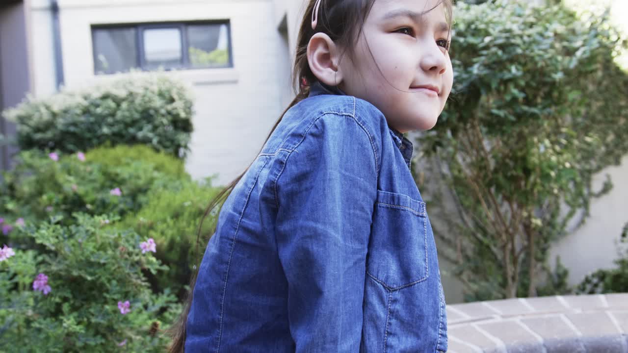カメラが上昇する中、レンガの縁に座ったデニム姿のアジア人の子供がラベンダー色のバッグを調整し、庭の方を見ている。