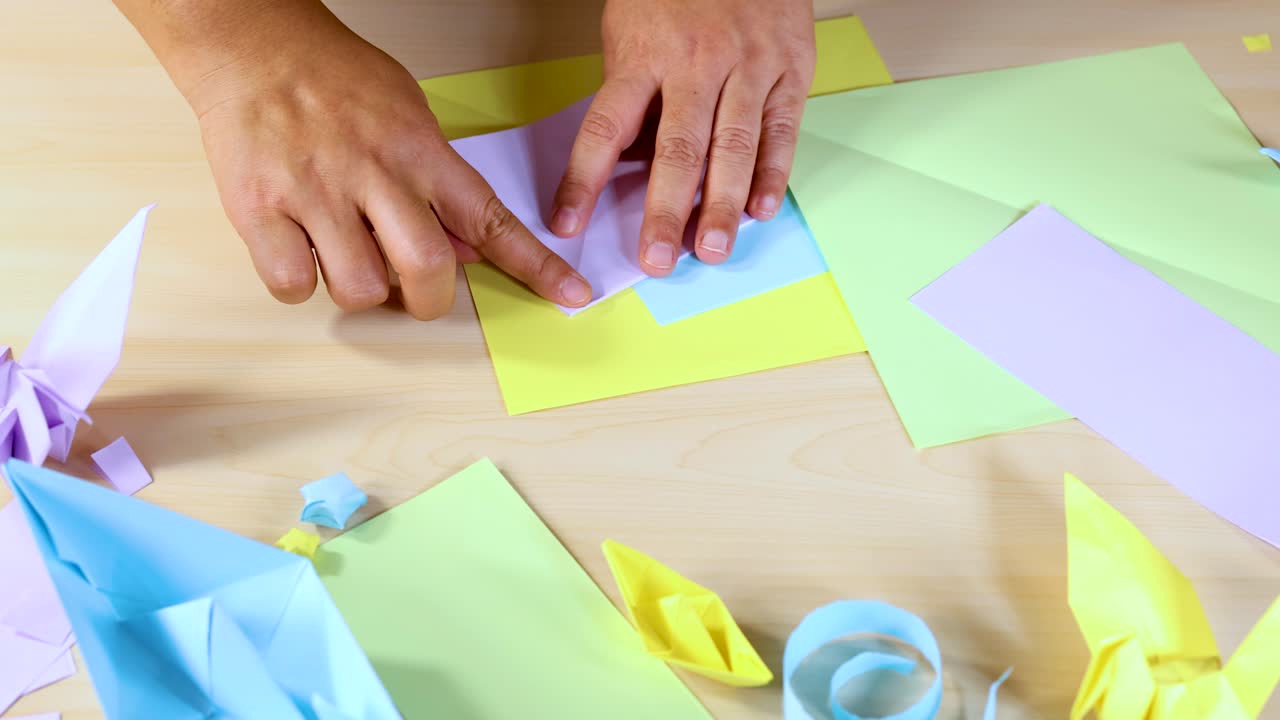 Person folds pastel origami paper, surrounded by completed paper crafts, under bright even lighting