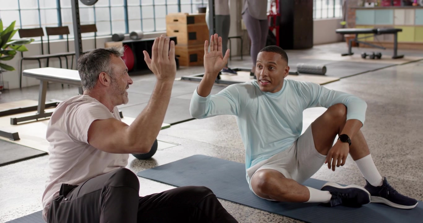Diverse male partners doing crunches on mats in gym after set cheering and giving high-five