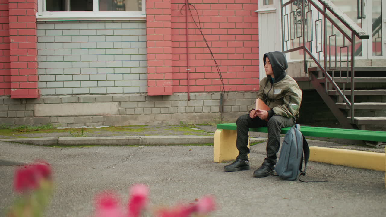 Teen in hooded jacket seated on outdoor bench holding book in right hand, backpack placed beside, tapping book thoughtfully, with blurred view of red flowers in foreground