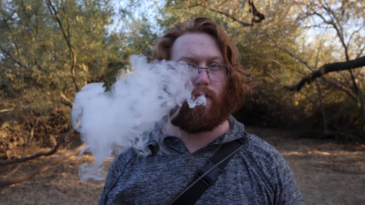 красноволосый мужчина показывает вэйп в камеру, а потом курит.