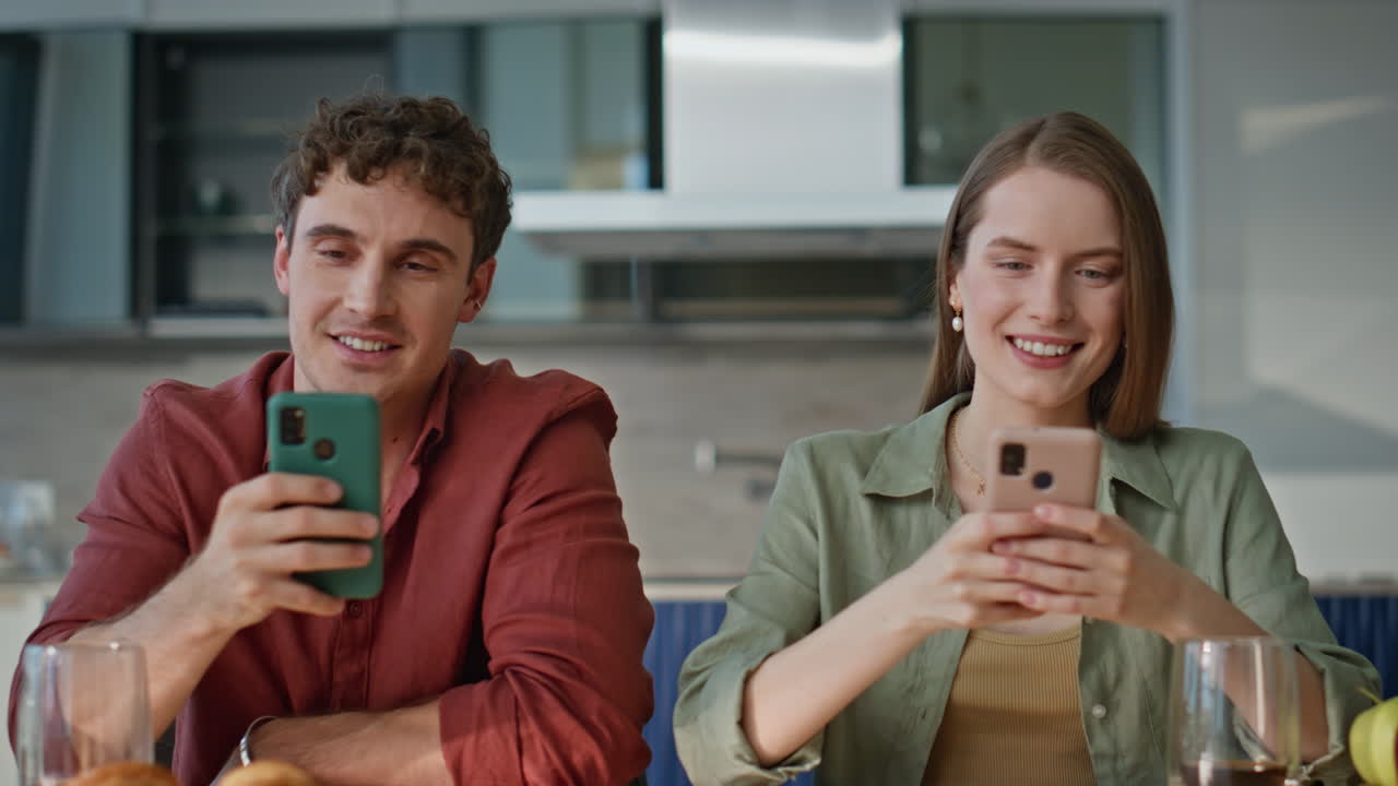 Cute spouses holding cellphones smiling to each other at modern kitchen closeup