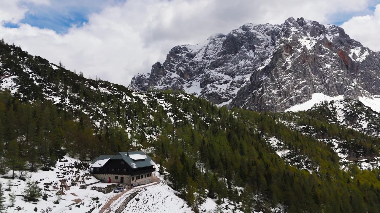 Il Rifugio del Postino sul Passo Vršič in Slovenia, circondato da foresta alpina, chiazze di neve e uno spettacolare paesaggio montano, vista aerea cinematografica.