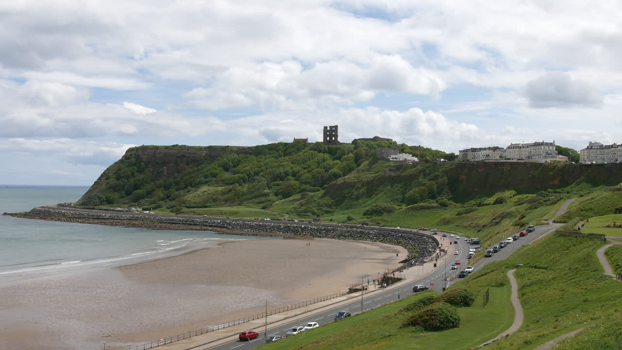 A castle ruin stands atop a hill surrounded by green trees, overlooking a wide sandy beach and a sea wall. Vehicles and people follow the road along the coast below the cliffs