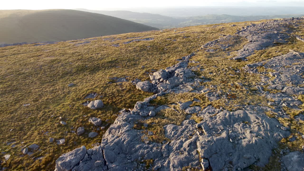 En 4K luftoptagelse med drone, der bevæger sig over et græsbeklædt bjerg med marker i baggrunden og solnedgang