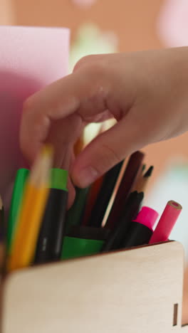 girl chooses colour pencil for drawing in wooden holder on table closeup. little student seeks crayon to paint image in classroom at art lesson