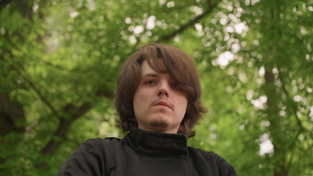 Close up of young man with shoulder length brown hair wearing black jacket seated outdoors in front of lush green trees. subject remains still maintaining direct gaze and neutral expression