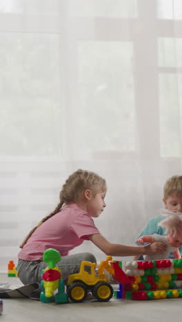 little boy watches elder sister shaking baby doll sitting on floor in living room. siblings enjoy playing with toys and constructor at home