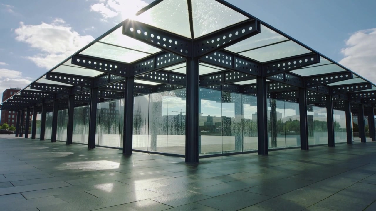La luz solar cambiante iluminando las columnas de un pabellón de vidrio y acero, y las estelas de agua brillando en una plaza frente al mar.