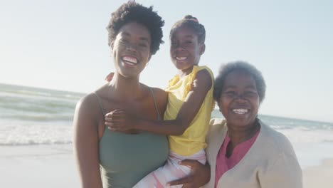 portrait of happy african american grandmother, mother and daughter smiling at beach, in slow motion