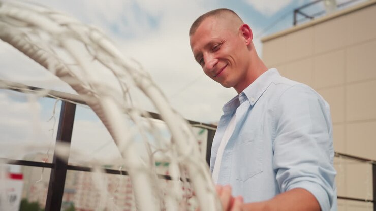 Uomo caucasico su un tetto che sorride mentre ispeziona un'altalena di vimini, camicia casual con bottoni, luce solare calda, ringhiera moderna della terrazza, atmosfera da appuntamento intimo, inquadratura cinematografica spontanea, profondità di campo ridotta