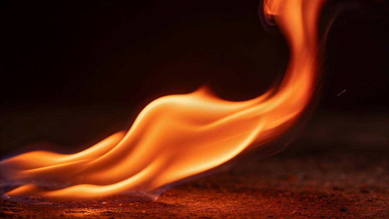 Sweeping bright orange flame ribbon moving across rough asphalt surface, small fuel pool igniting