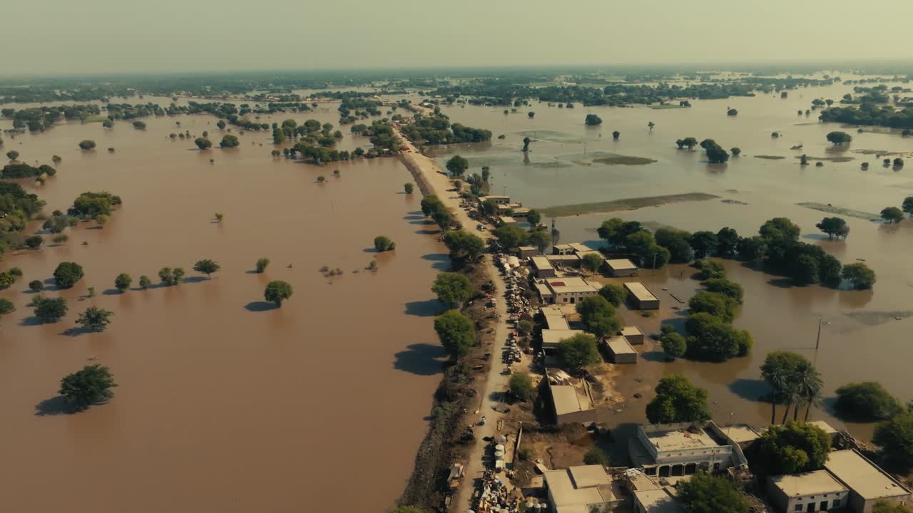 Drone flight over dry road surrounded by flood waters, showing people walking, emergency tents, and houses on a sunny day
