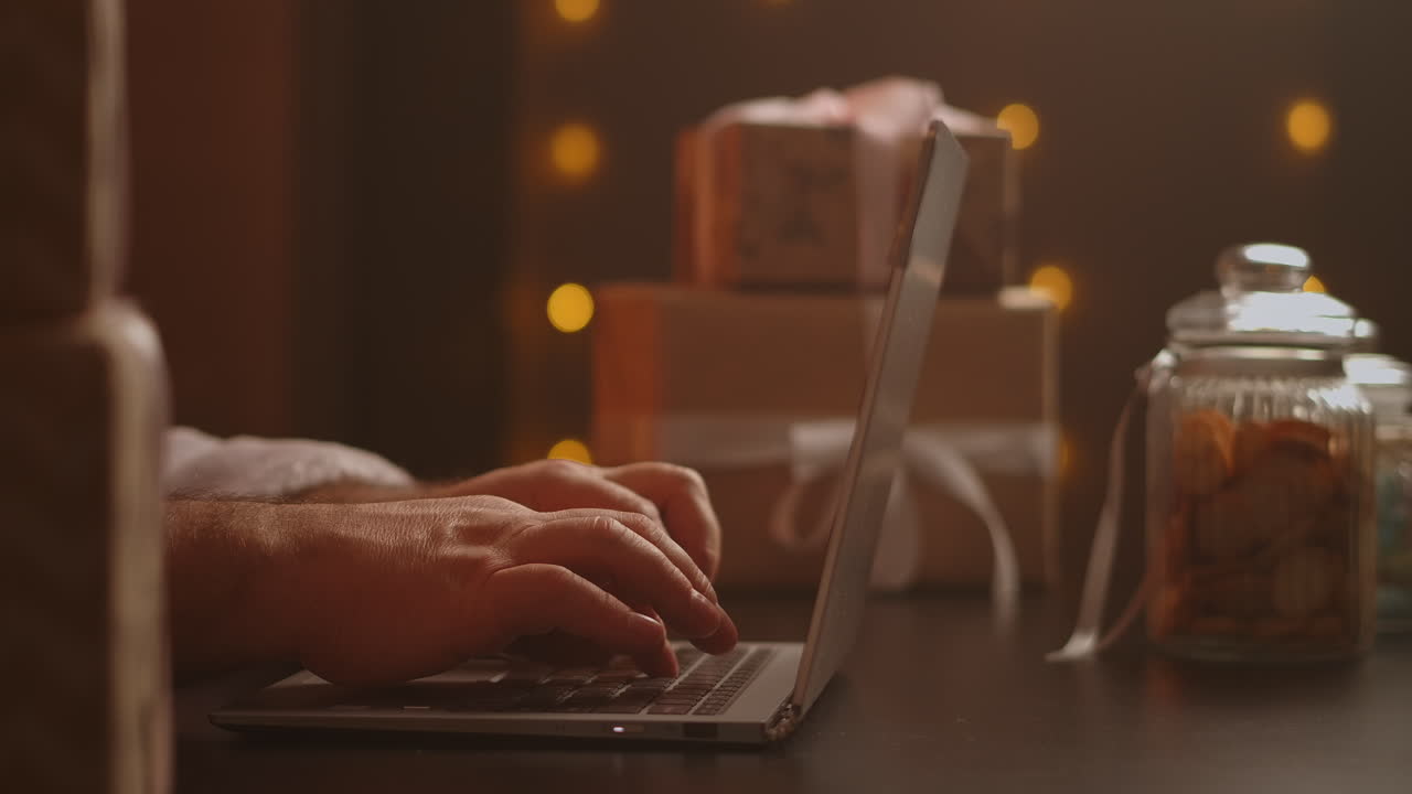Handen van de Kerstman typen op een draadloos toetsenbord bij een houten, voor Nieuwjaar versierde tafel. De Kerstman werkt met een laptop, kijkt door zijn mail en beantwoordt berichten aan kinderen. Hoge kwaliteit 4K-beelden