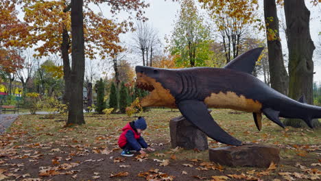 caucasian toddler standing near the wooden sculpture of shark. kid picks up the acorns and puts in shark jaws.