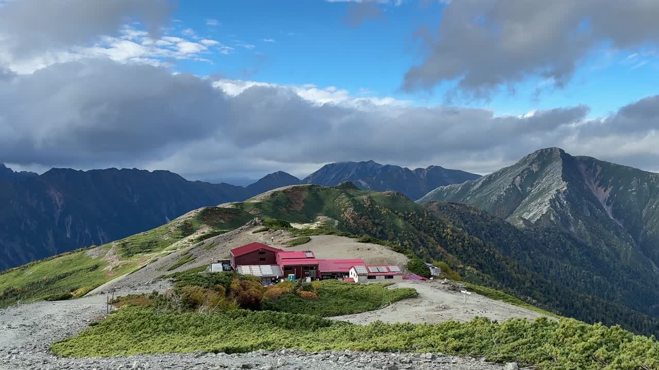 Wide shot of Chogatake Hut on an alpine ridge with the Hotaka mountains rising behind it under a bright, dramatic sky