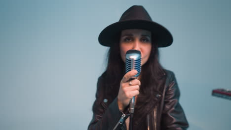 a vocalist in a black leather jacket and hat singing with eyes open,stands at a vintage microphone held with one hand. a keyboardist plays sampler against a blue background as the camera zooms