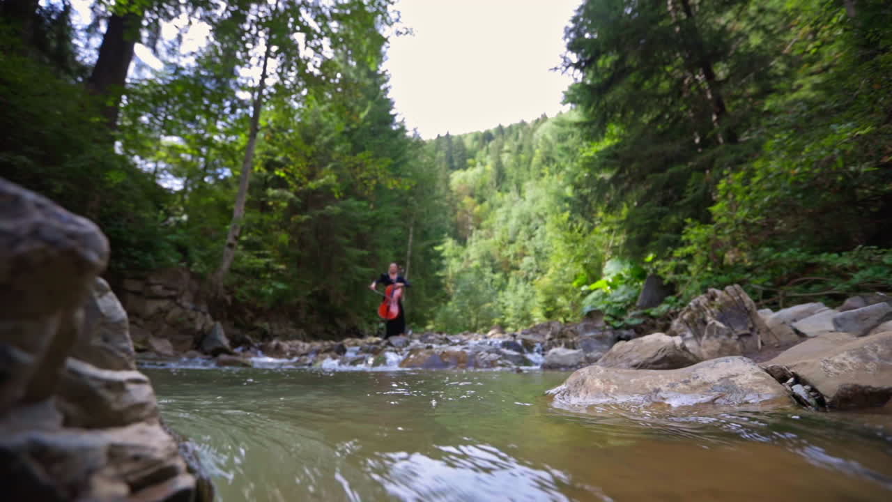 Cellist among mountain water. Professional female musician performing music with large musical instrument on green nature background.