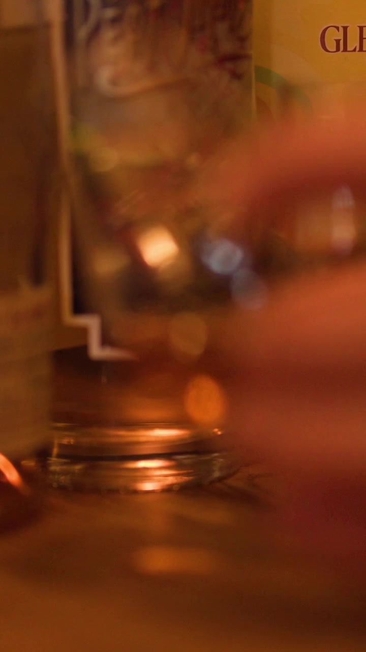 Una mano sfocata cerca un bicchiere di whisky ambrato sotto la calda luce di un pub