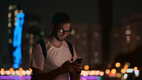 Ein-Männlicher-Reisender-Mit-Rucksack-Und-Brille-Vor-Dem-Hintergrund-Der-Nächtlichen-Stadt-Ruft-über-Ein-Mobiltelefon-Ein-Taxi.-Hält-Ein-Smartphone-Und-Berührt-Den-Bildschirm.-Sieht-Sich-Fotos-An-Und-Schreibt-Nachrichten-In-Sozialen-Netzwerken