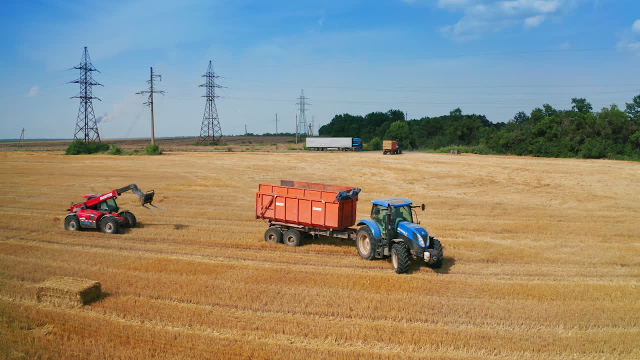 Big empty tractor moves by the mowed field. Vast farmlands of harvested wheat with hay bales at backdrop.