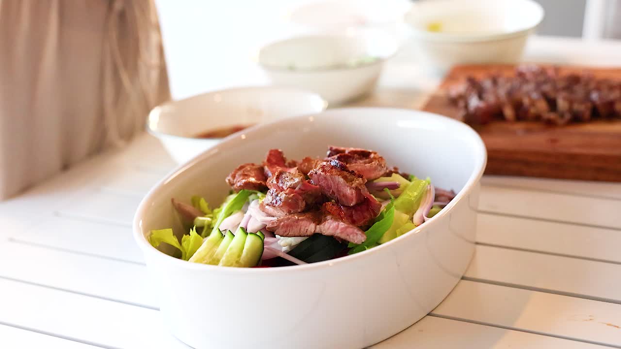 Hands arrange beef on salad in bright kitchen. Natural lighting highlights fresh ingredients and vibrant colors