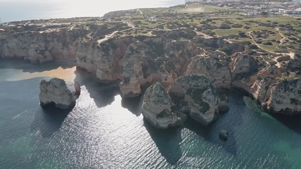 Cinematic aerial orbit shot where the drone circles the iconic golden cliffs of Lagos, Portugal as a boat transporting kayaks glides smoothly through turquoise waters beneath