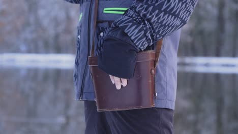 a person in a blue jacket standing on a snowy riverbank has his hand on a leather case for binoculars