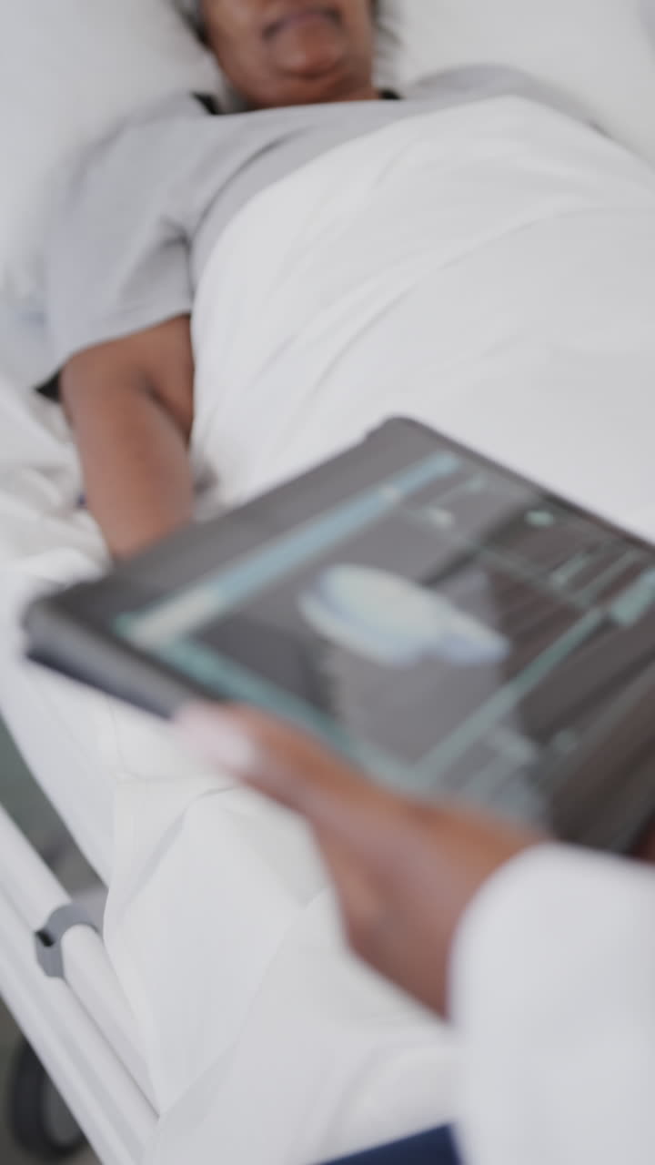 Vertical video of diverse female doctor with tablet and senior female patient on drip, slow motion