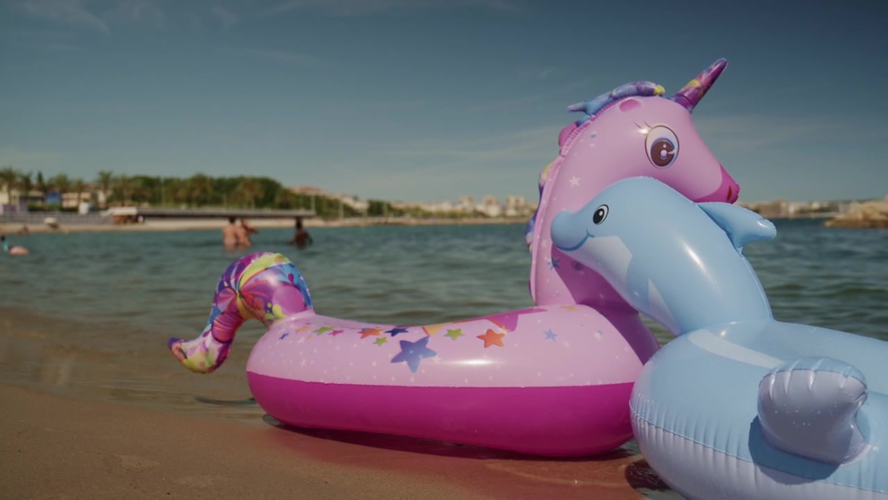 Antibes, França - 4 de junho de 2026: Boias infláveis coloridas de unicórnio e golfinho em águas rasas na Plage de la Gravette com famílias nadando e o quebra-mar rochoso ao fundo.