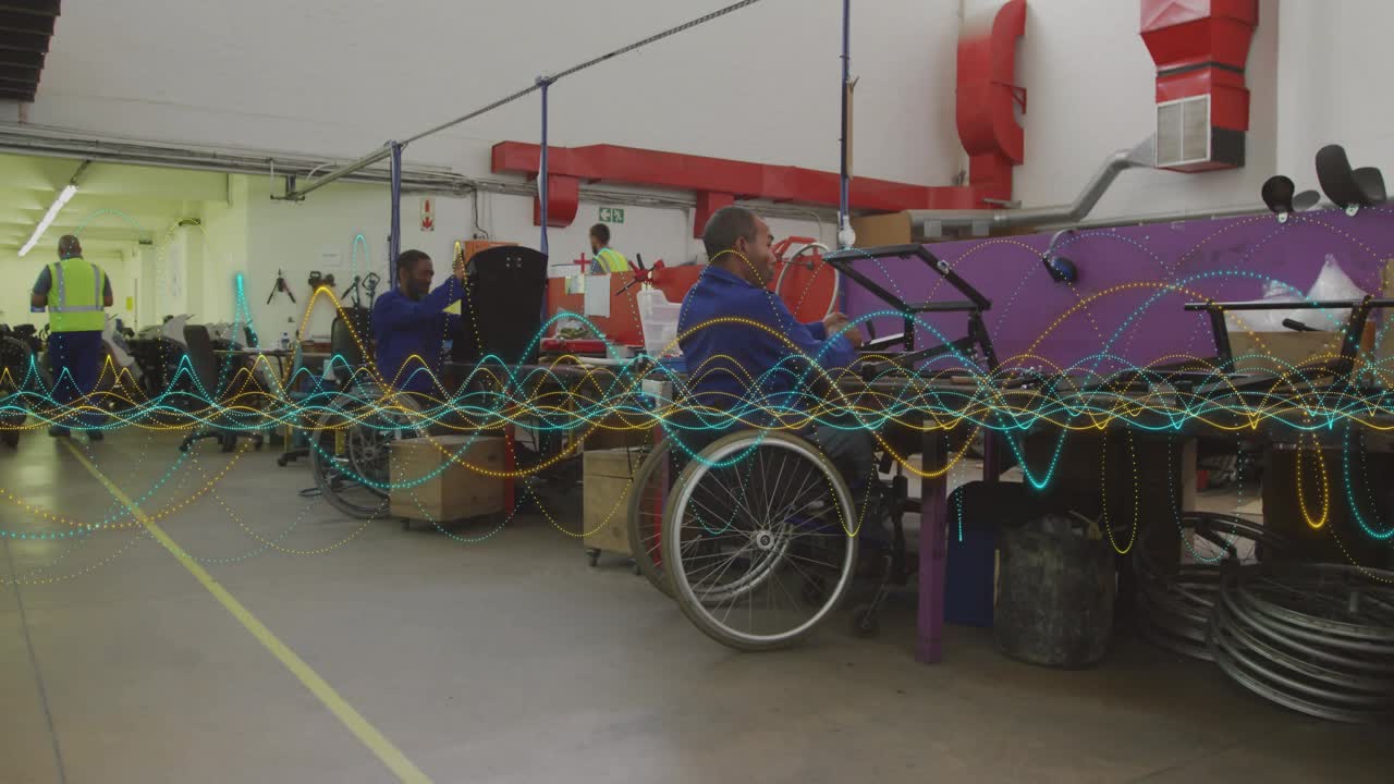 Man seated in manual wheelchair working at purple bench in workshop, passing workers obscuring view