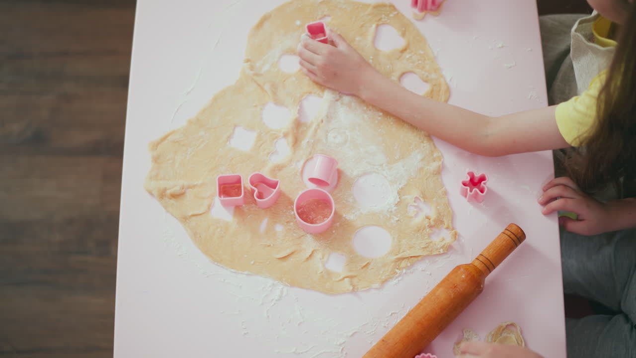 Top down view of girl placing flour into pink shape cutter while baking, with another kid busy working on dough, surrounded by baking tools, cookie cutters, and scattered flour on table